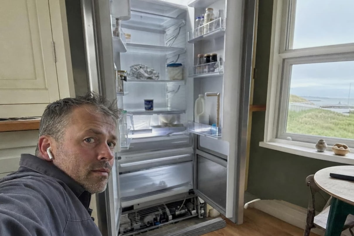 Technician beside an open built-in refrigerator in a coastal kitchen, the lower machinery compartment open at the floor and water visible through the window