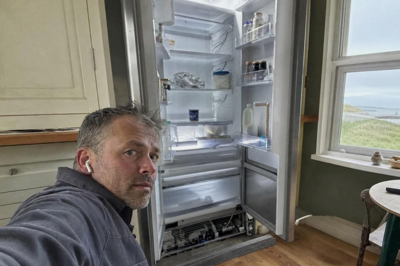 Technician kneeling beside an open built-in refrigerator in a coastal kitchen, the lower grille off and the machinery visible