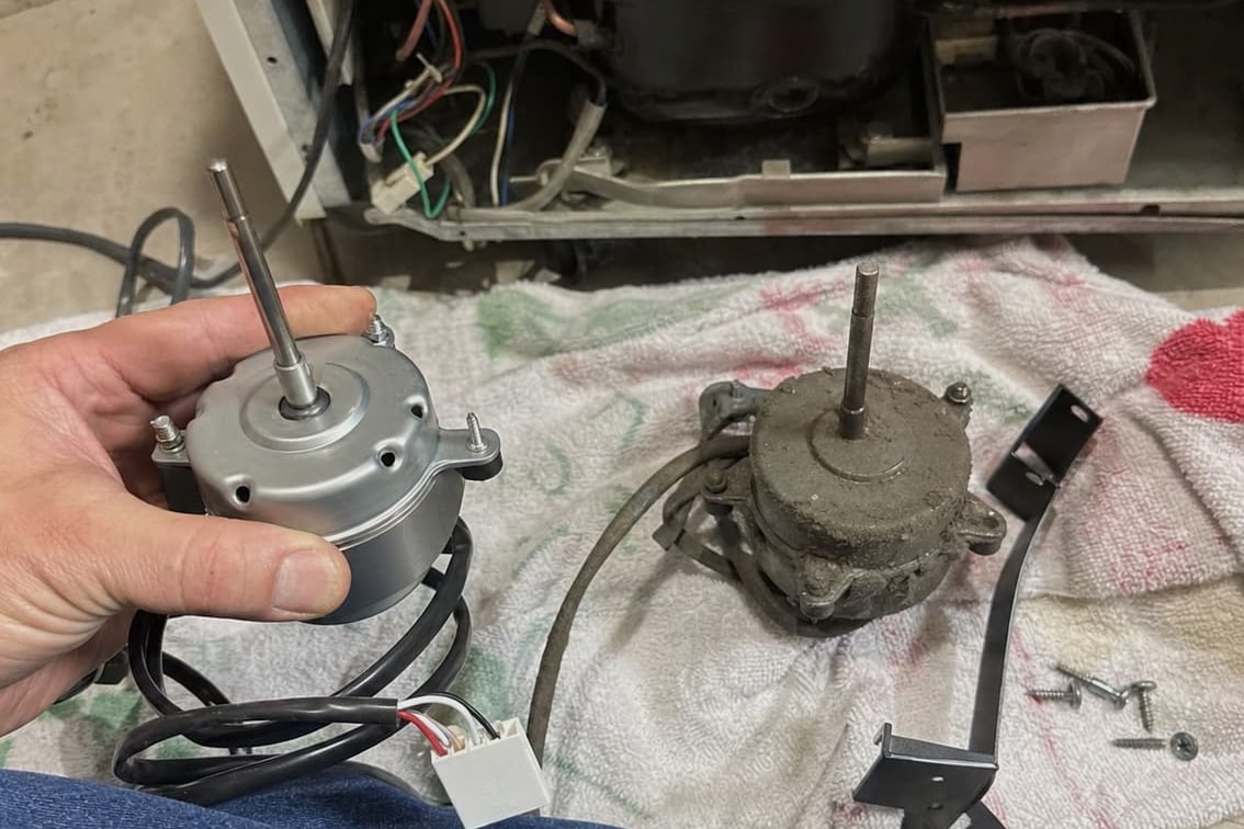 New condenser fan motor held beside the corroded one it is replacing, with the compressor bay open behind