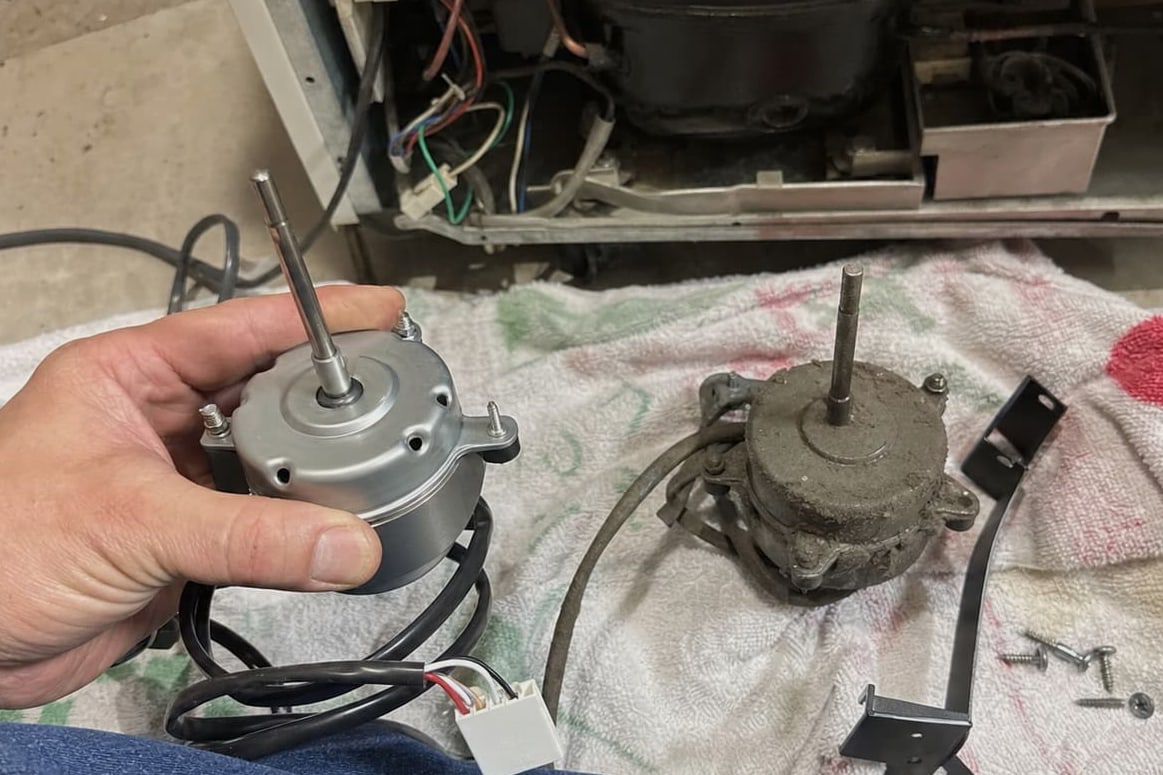 New condenser fan motor held beside the corroded one it is replacing, with the compressor bay open behind