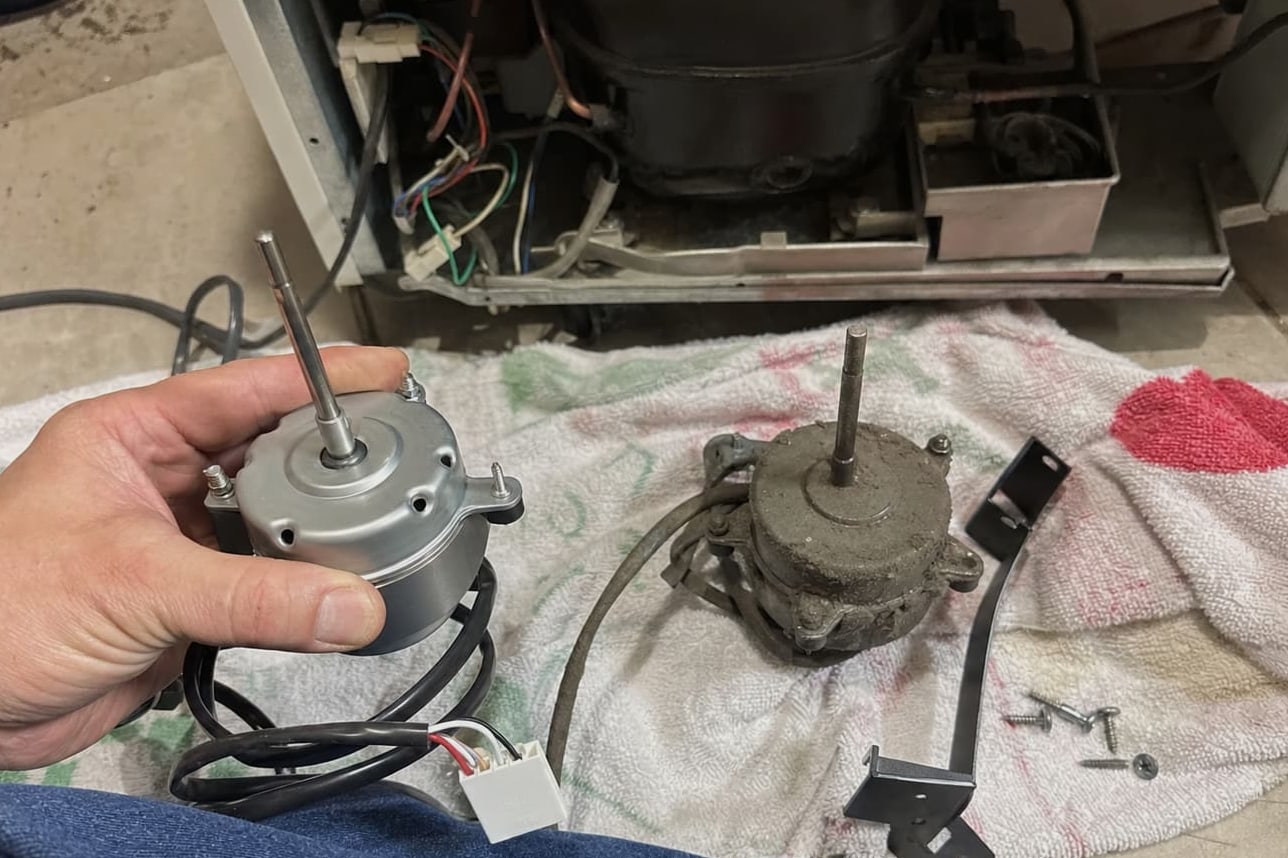 New condenser fan motor held beside the corroded one it is replacing, with the compressor bay open behind