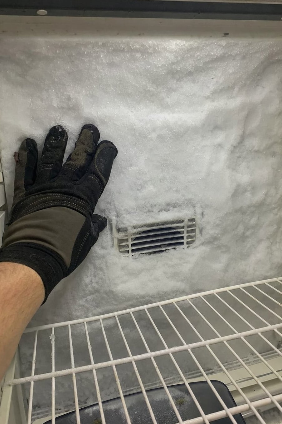 Gloved hand against a thick frost wall on the rear panel of a freezer compartment, the air vent buried in ice