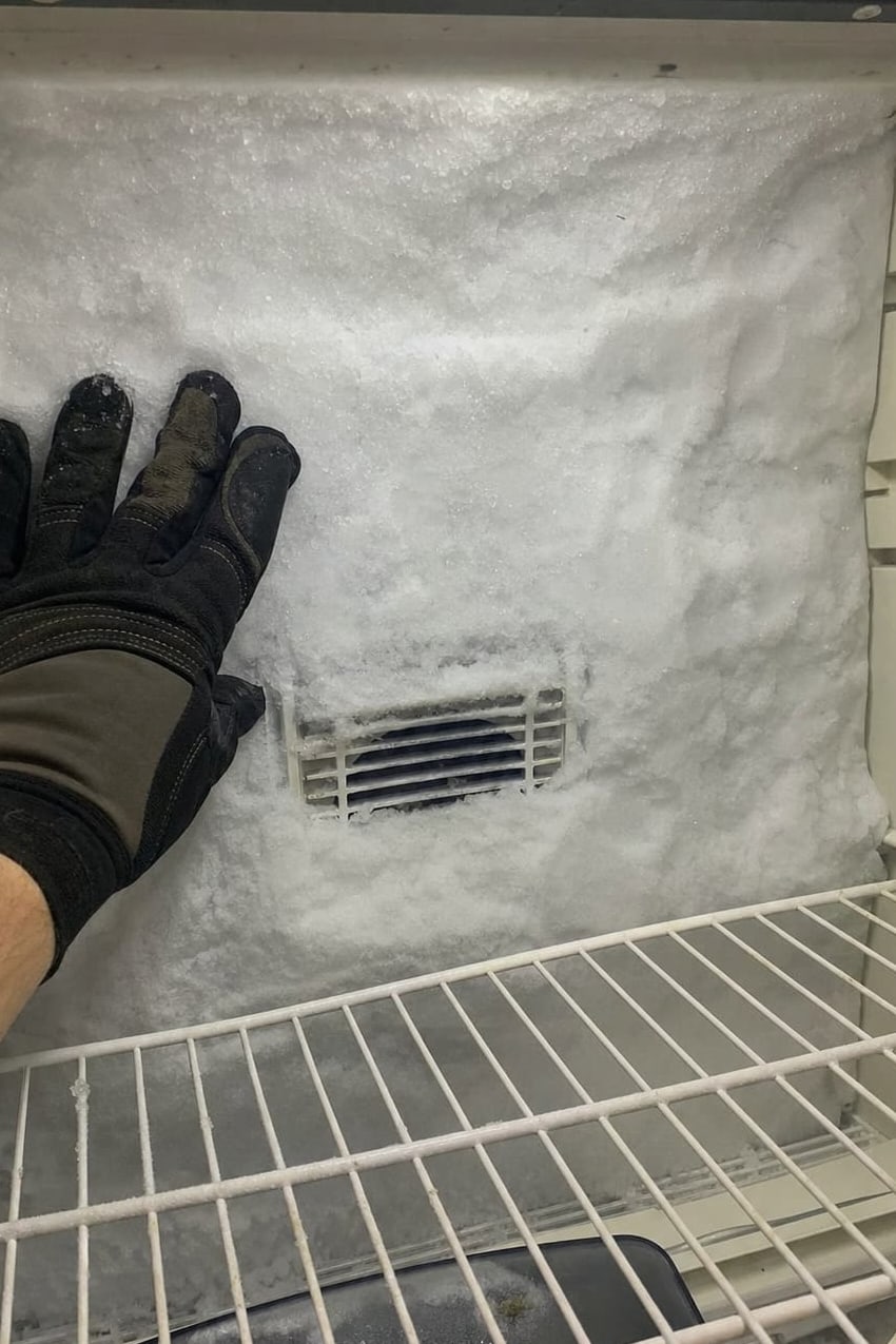 Gloved hand against a thick frost wall on the rear panel of a freezer compartment, the air vent buried in ice