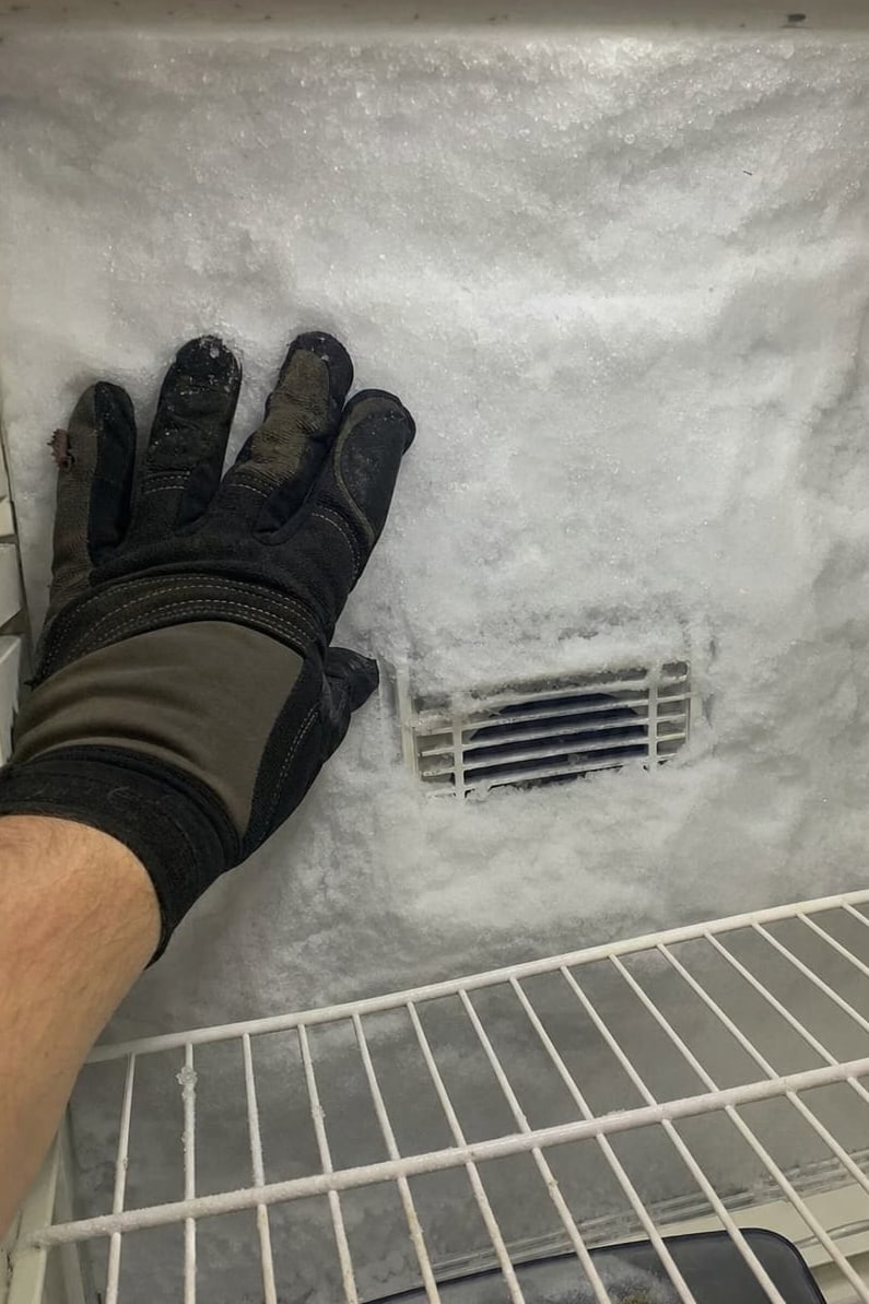 Gloved hand against a thick frost wall on the rear panel of a freezer compartment, the air vent buried in ice