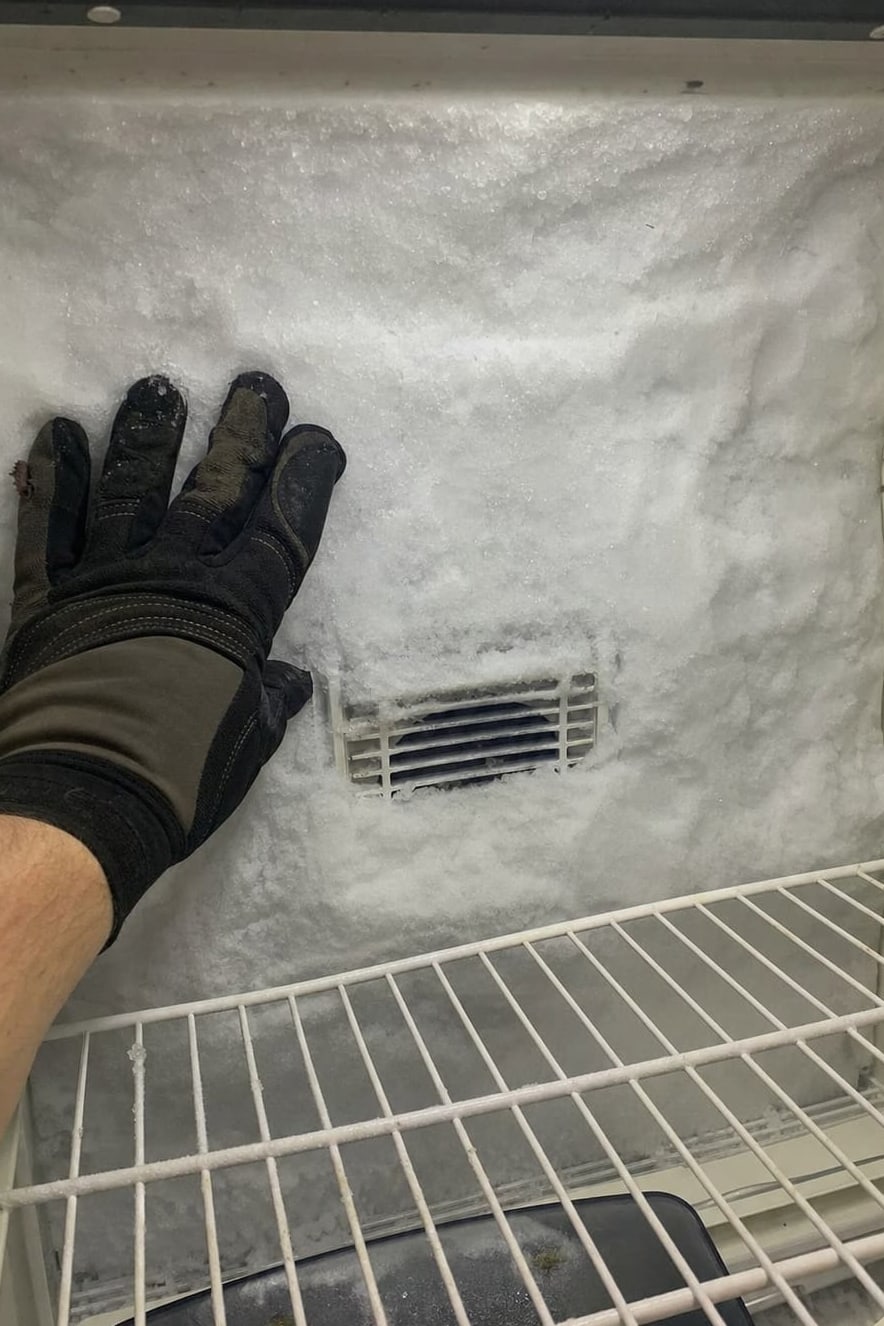 Gloved hand against a thick frost wall on the rear panel of a freezer compartment, the air vent buried in ice
