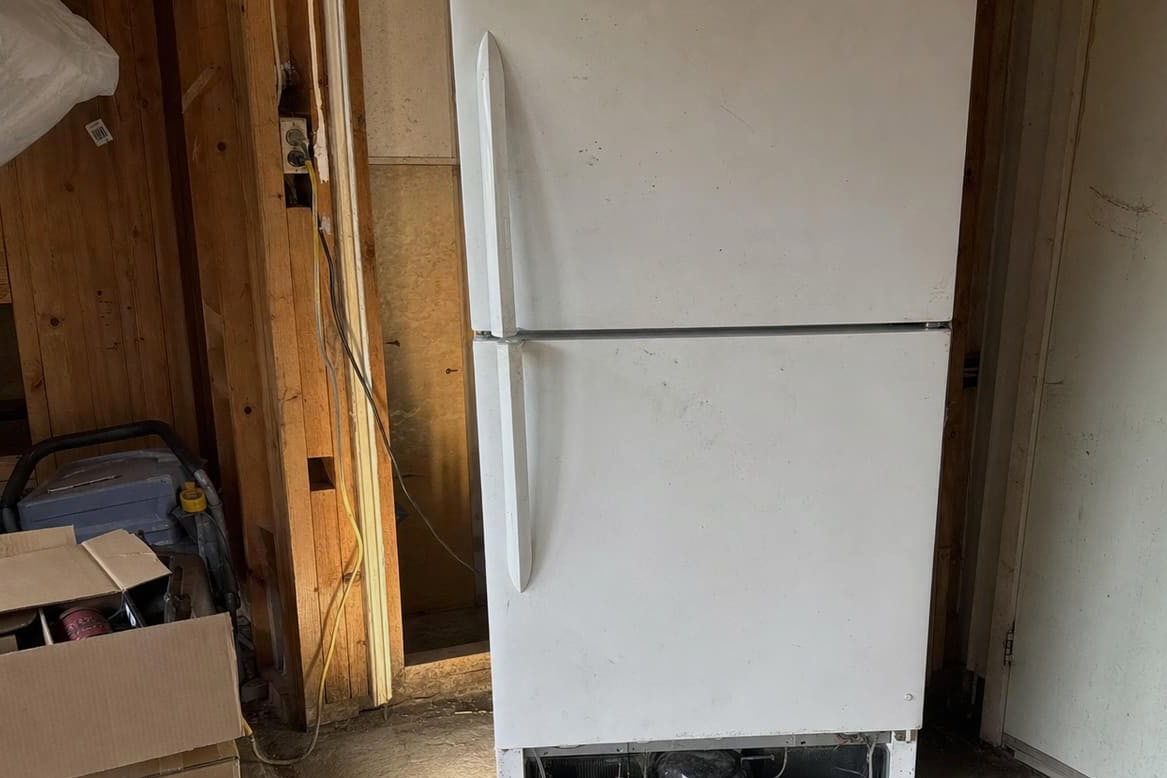 White top-freezer refrigerator standing against the bare timber wall of an unconditioned garage, cardboard boxes and stored gear beside it