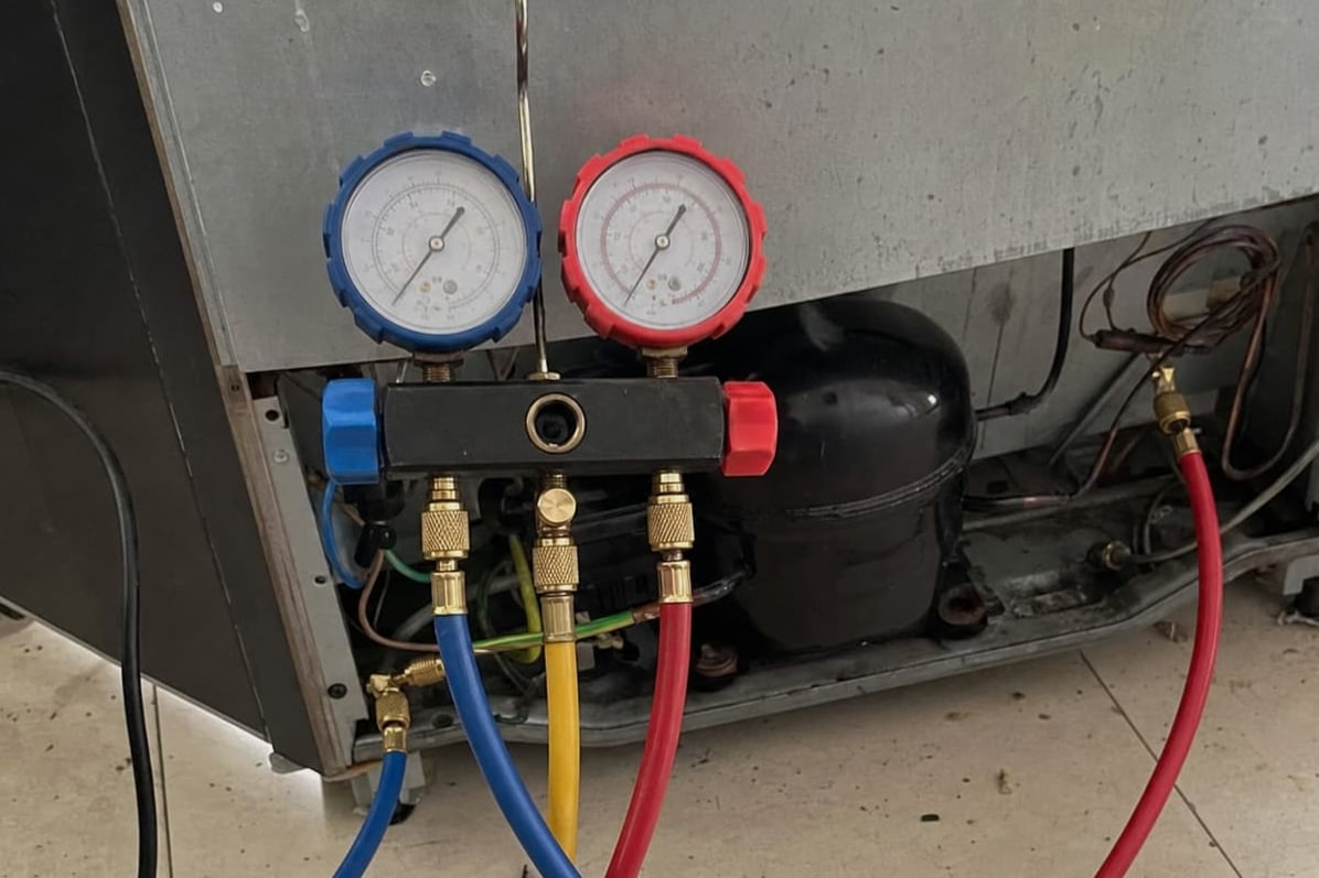 Manifold gauge set connected at the compressor behind a refrigerator, blue, yellow and red hoses running to the service ports