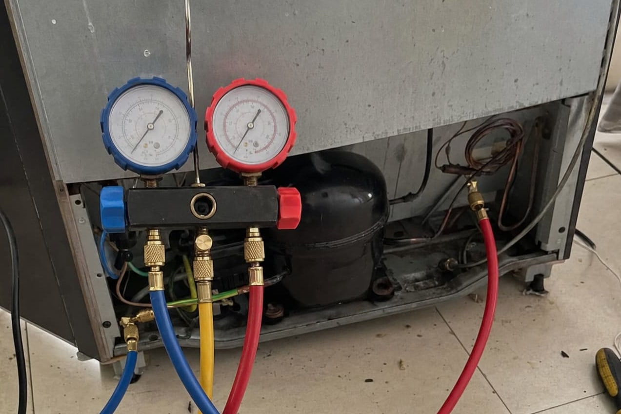 Manifold gauge set connected at the compressor behind a refrigerator, blue, yellow and red hoses running to the service ports