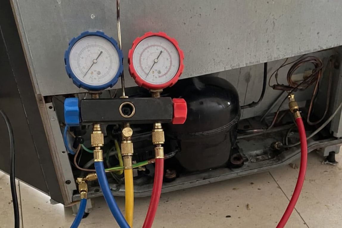 Manifold gauge set connected at the compressor behind a refrigerator, blue, yellow and red hoses running to the service ports