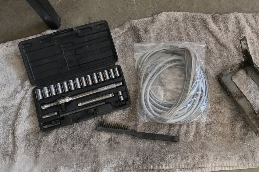 Socket set, wire brush, coiled charging hoses and a grimy condenser bracket laid out on a towel on a garage floor