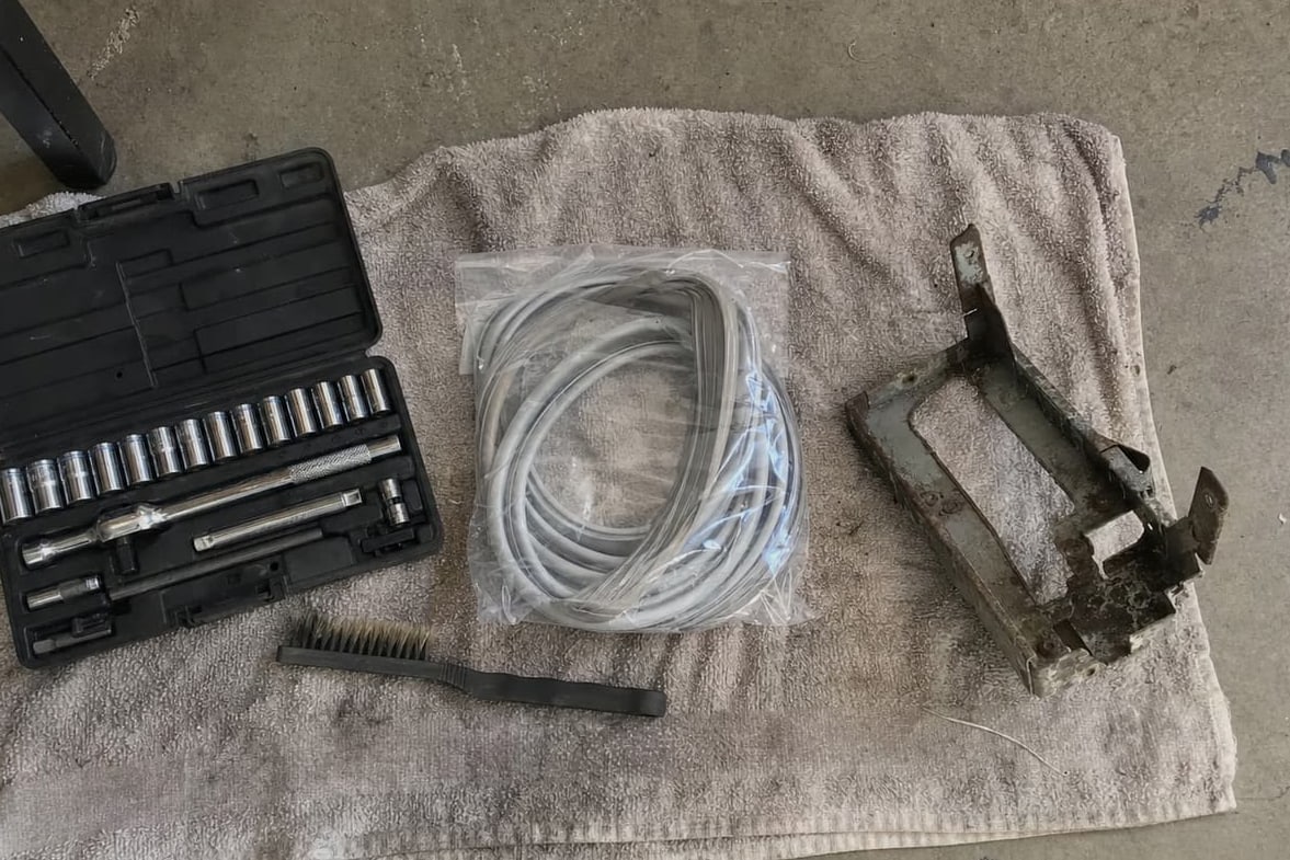 Socket set, wire brush, coiled charging hoses and a grimy condenser bracket laid out on a towel on a garage floor