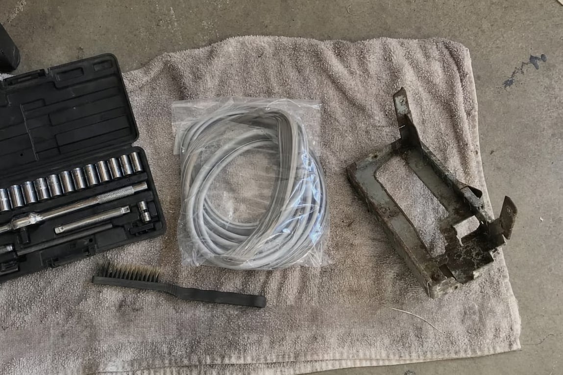 Socket set, wire brush, coiled charging hoses and a grimy condenser bracket laid out on a towel on a garage floor