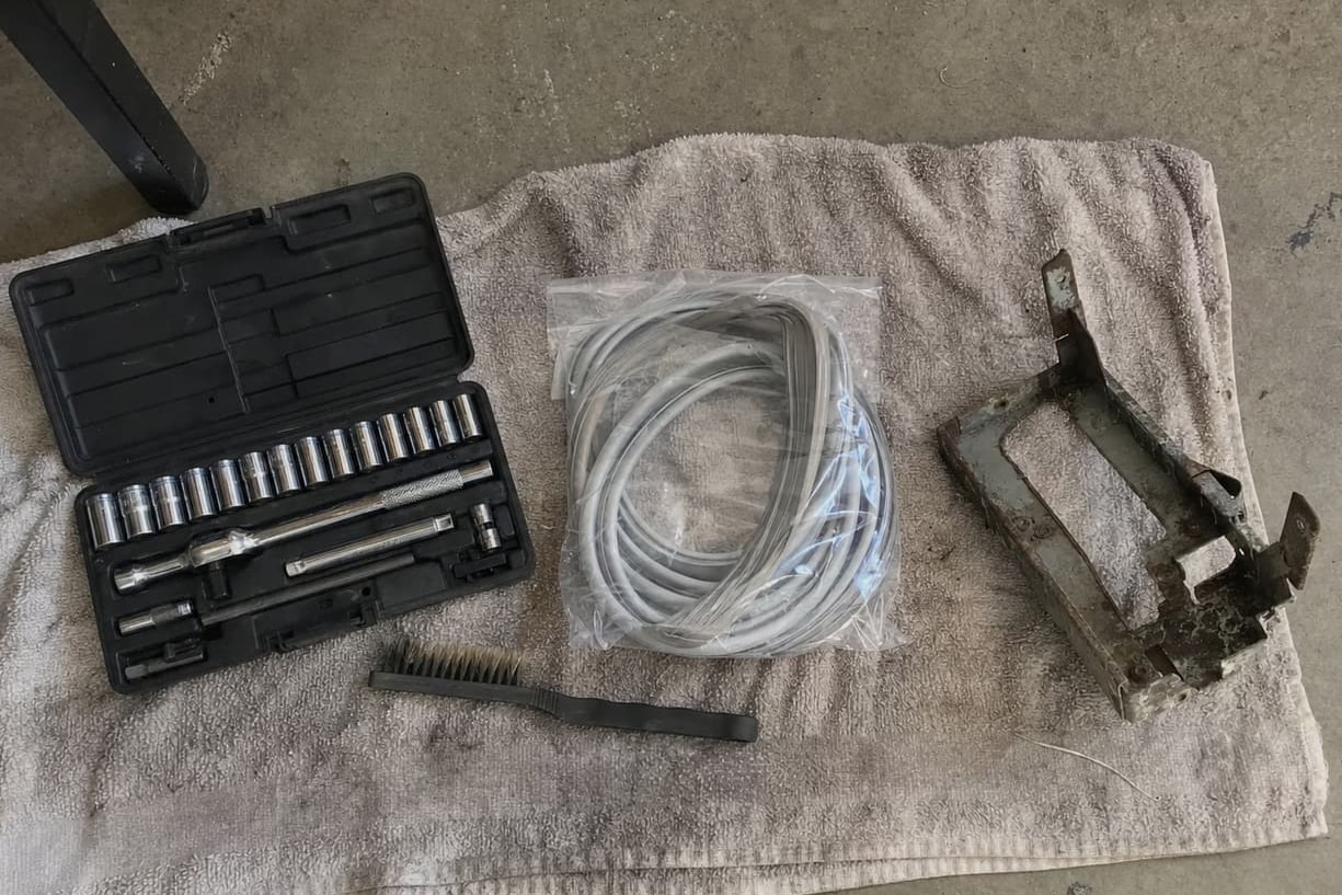 Socket set, wire brush, coiled charging hoses and a grimy condenser bracket laid out on a towel on a garage floor