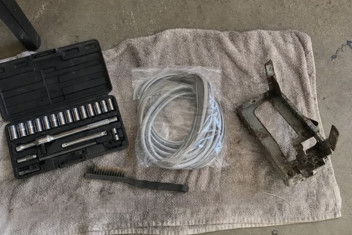 Socket set, wire brush, coiled charging hoses and a grimy condenser bracket laid out on a towel on a garage floor