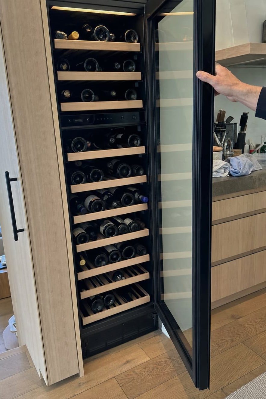 Wine storage column standing open in a kitchen, full racks of bottles on light wooden shelves, a hand holding the glass door back